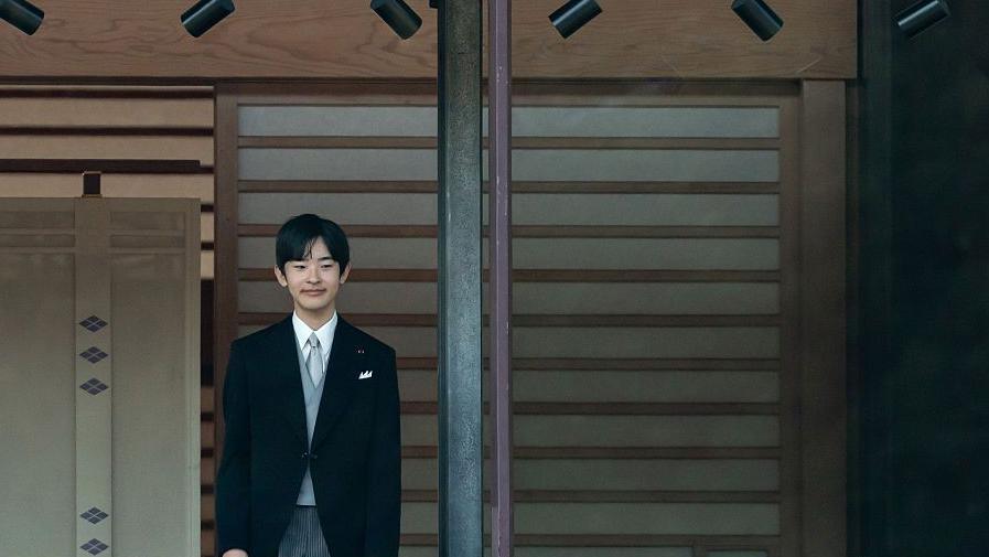 เจ้าชายฮิซาฮิโตะแย้มสรวลและประทับยืนหลังหน้าต่าง พระองค์ทรงฉลองพระองค์สูทสีดำ