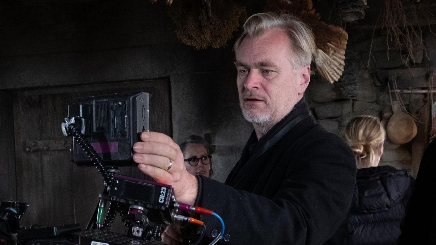 คริสโตเฟอร์ โนแลน อยู่กลางภาพและกำลังมองผ่านกล้อง Imax เขาสวมใส่แจ็คเก็ตสีดำ