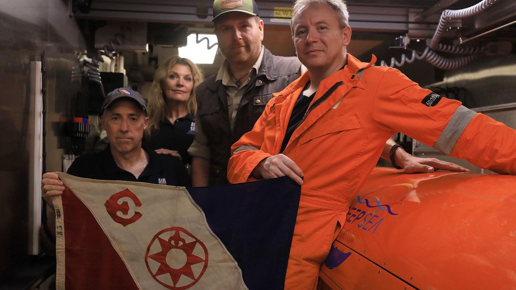 ภาพ Russ Matthews ถือธง Explorers Club กับสมาชิกทีมของเขาและ Josh Gates