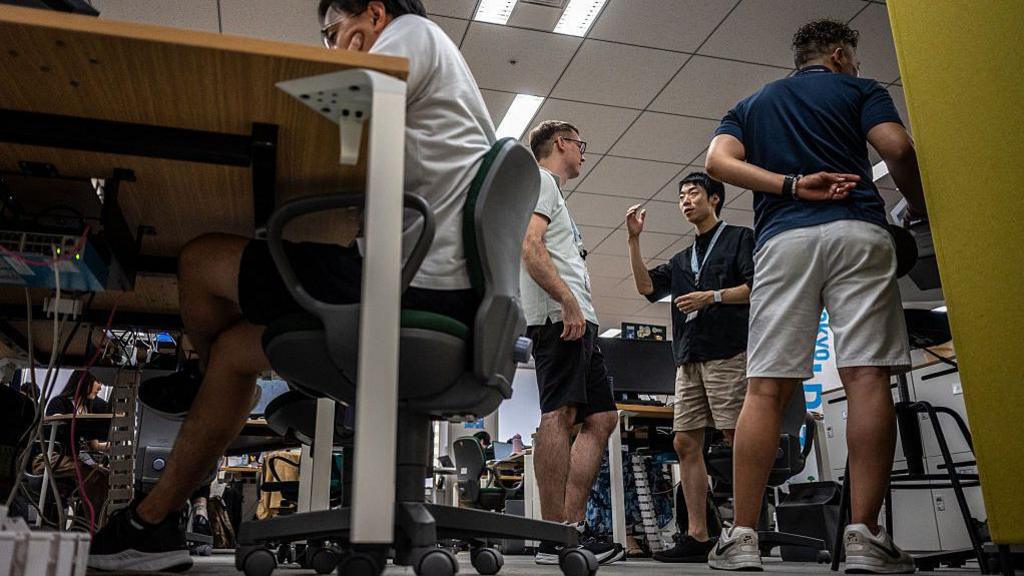 ชายสี่คนสวมกางเกงขาสั้นในสำนักงานที่กรุงโตเกียว ประเทศญี่ปุ่น โดยสามคนกำลังยืน และหนึ่งคนนั่งโดยวางข้อศอกบนโต๊ะ