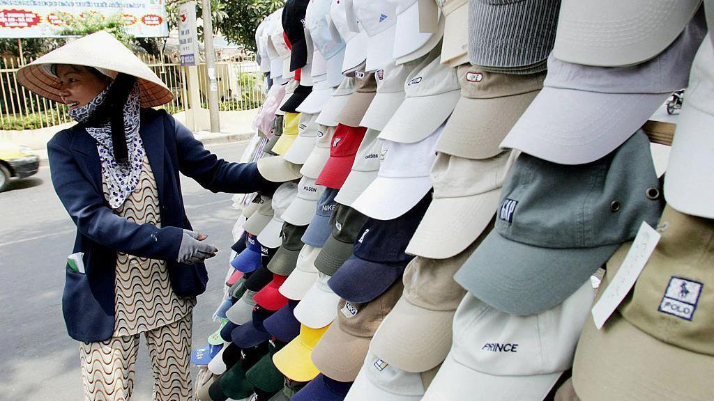 หมวกที่มีโลโก้ Polo, GAP, Prince และอื่นๆ แขวนอยู่เบื้องหน้า ถัดจากผู้หญิงที่สวมเสื้อเบลเซอร์สีน้ำเงินและหมวกงอนละ หรือหมวกใบไม้ฟาง