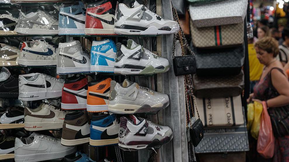 รองเท้าผ้าใบ Nike ที่ห่อด้วยพลาสติกวางเรียงรายอยู่ในตลาดข้างกระเป๋าถือ ขณะที่ผู้คนกำลังเลือกซื้อสินค้าอยู่เบื้องหลัง