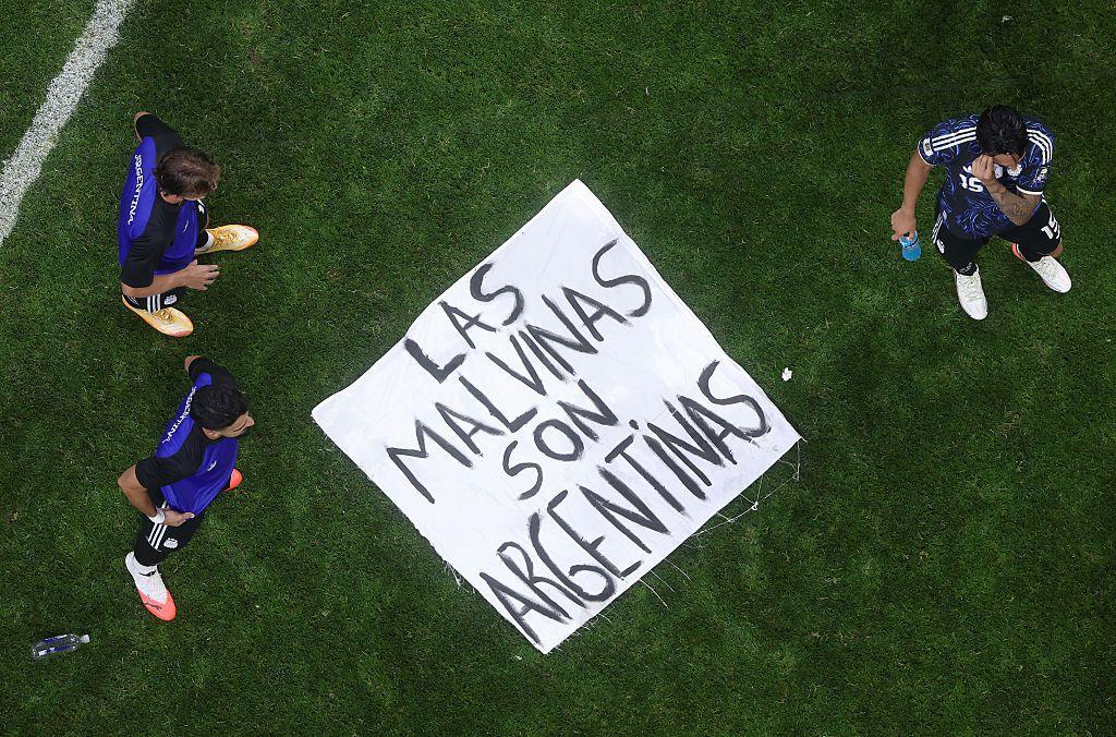 ป้ายที่มีข้อความว่า "Las Malvinas Son Argentinas" วางอยู่บนพื้นสนาม โดยมีผู้เล่นสามคนยืนอยู่รอบๆ