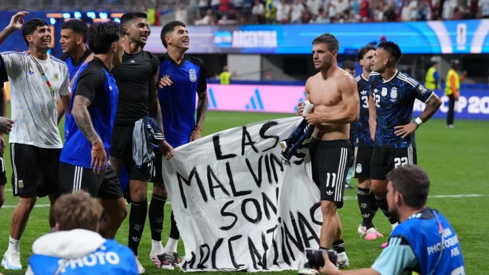 นักฟุตบอลอาร์เจนตินาหลายคนยืนอยู่บนสนามพร้อมชูป้ายที่มีข้อความว่า "Las Malvinas Son Argentinas"