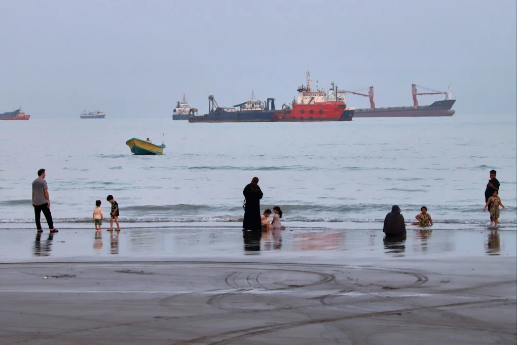 ผู้คนพักผ่อนอยู่บนชายหาด ในขณะที่เรือพาณิชย์จอดทอดสมออยู่ในช่องแคบฮอร์มุซ นอกชายฝั่งบันดาร์อับบาส ประเทศอิหร่าน เมื่อวันพุธ