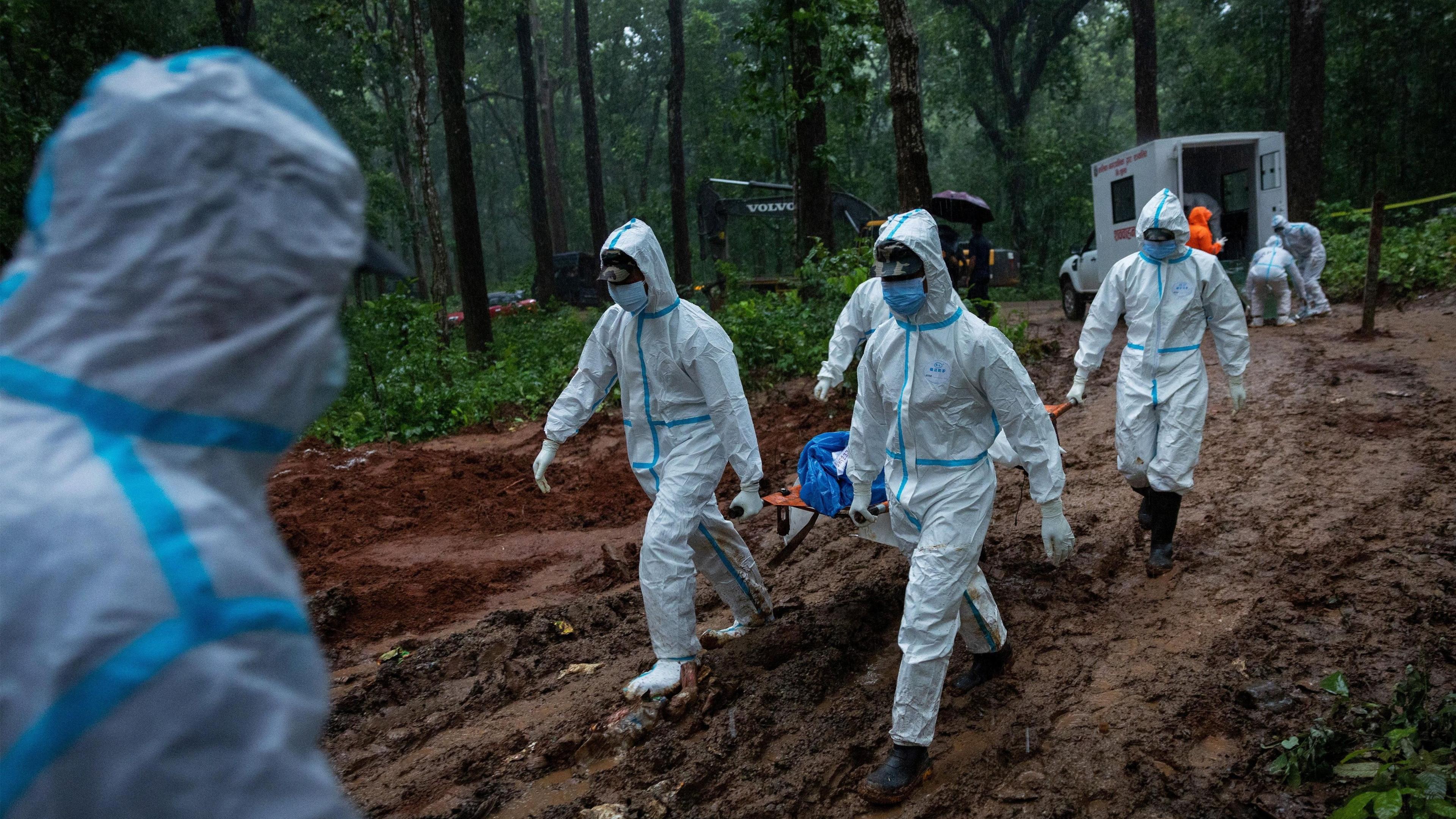ผู้คนในชุด PPE สีขาวกำลังแบกเปลที่มีศพอยู่บนนั้นในป่า