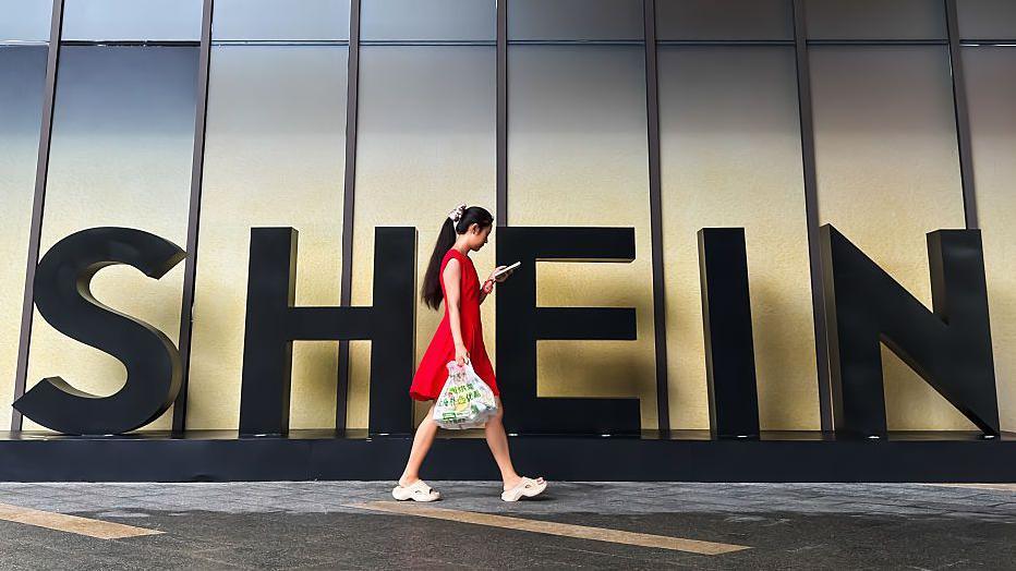 ผู้หญิงในชุดสีแดงมองสมาร์ทโฟนขณะเดินผ่านป้าย Shein นอกสำนักงานบริษัทในกว่างโจว ประเทศจีน