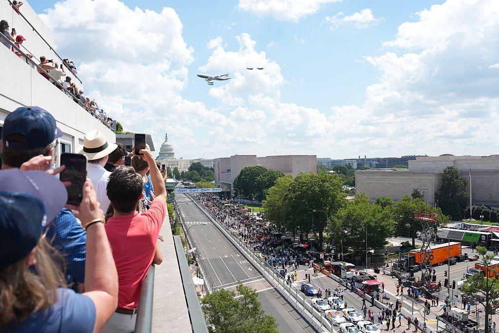 ผู้คนถ่ายภาพบนหลังคาขณะเครื่องบิน Air Force One บินโฉบโดยมีอาคารรัฐสภาอยู่เบื้องหลังและสนามแข่งอยู่ด้านล่าง