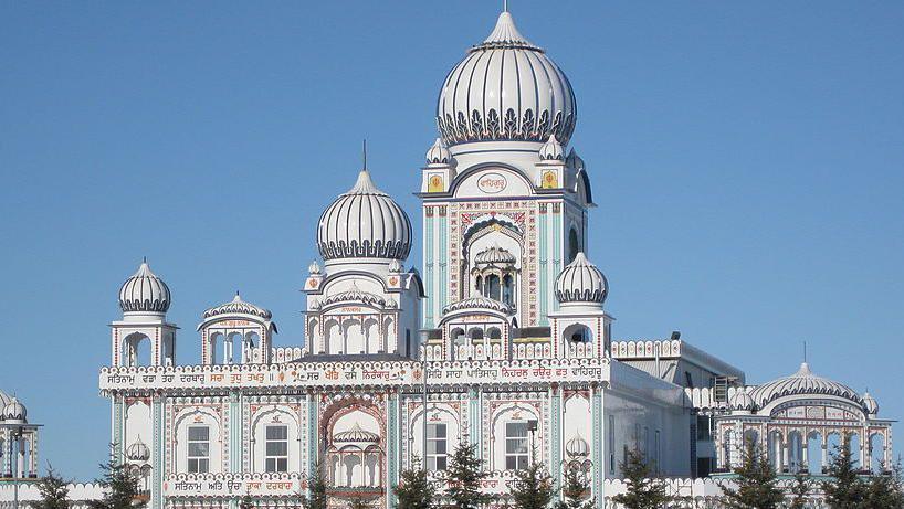 ภาพด้านนอกของโบสถ์ Nanaksar Gurdwara Gursikh Temple ในเมืองเอ็ดมันตัน เป็นอาคารขนาดใหญ่สีขาววิจิตรงดงาม ตกแต่งด้วยสีฟ้าอ่อนและสีกุหลาบ มีซุ้มโค้งและหลังคาทรงโดมหลายหลัง ล้อมรอบด้วยต้นสน