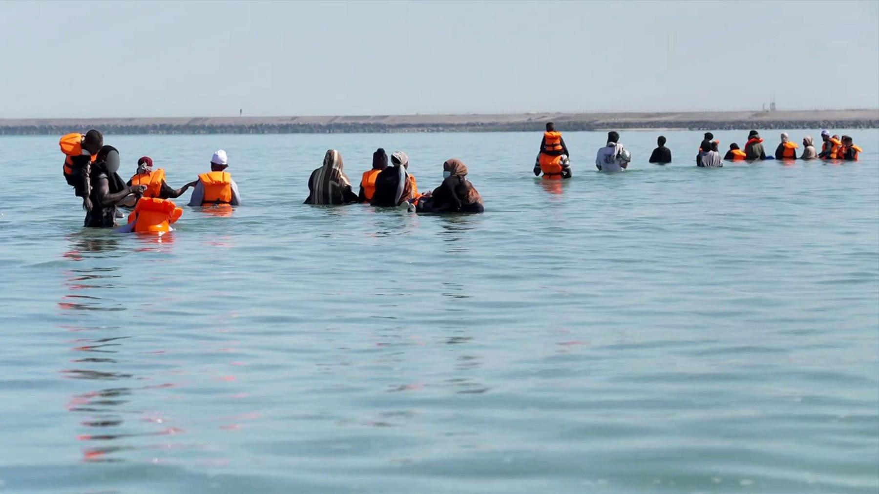 กลุ่มผู้อพยพเดินลุยน้ำทะเลบนชายหาดในกราฟไลน์ส