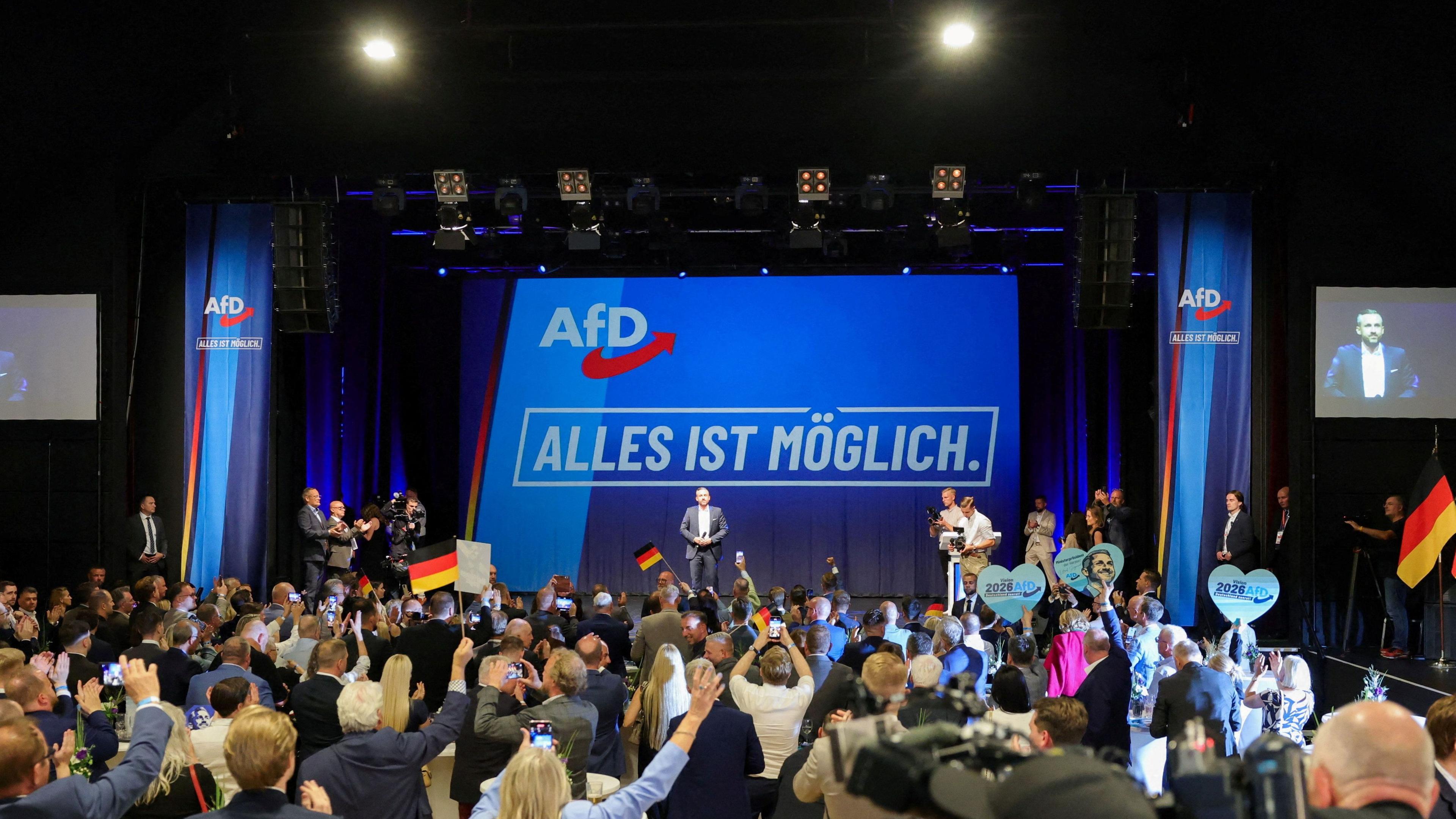 ฝูงชนโบกธงชาติเยอรมนีหน้าเวทีที่มีพื้นหลังสีน้ำเงินและโลโก้ของพรรค AfD