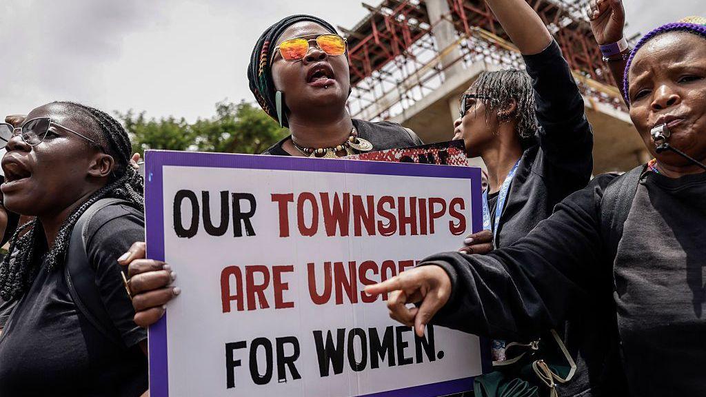 ผู้หญิงในการประท้วงสวมชุดสีดำ หนึ่งคนถือป้ายเขียนว่า: "Our townships are unsafe for women" (เมืองของเราไม่ปลอดภัยสำหรับผู้หญิง)