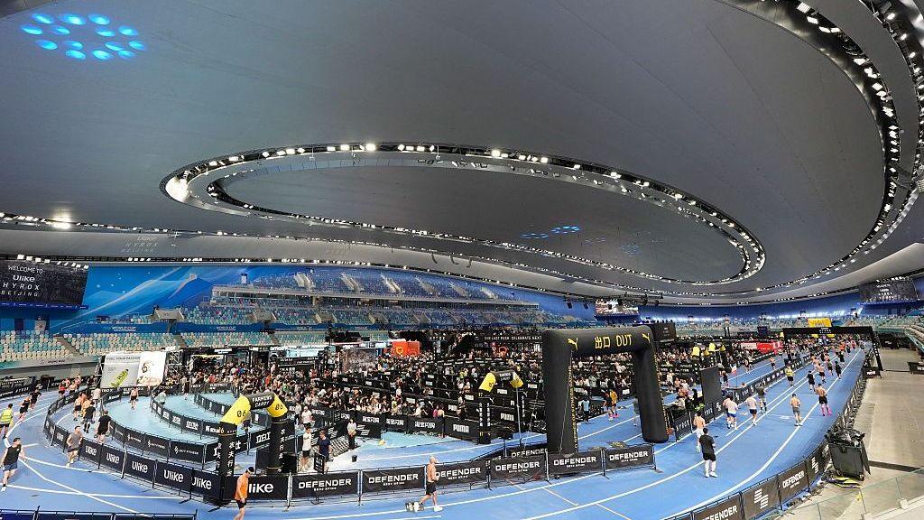 มุมมองจากอัฒจันทร์ของสนามกีฬา National Speed Skating Oval ในกรุงปักกิ่ง ซึ่งเป็นสถานที่จัดงาน Hyrox มีนักกีฬาจำนวนมากกำลังวิ่งอยู่รอบสนาม โดยมีสิ่งกีดขวางกั้นเลน