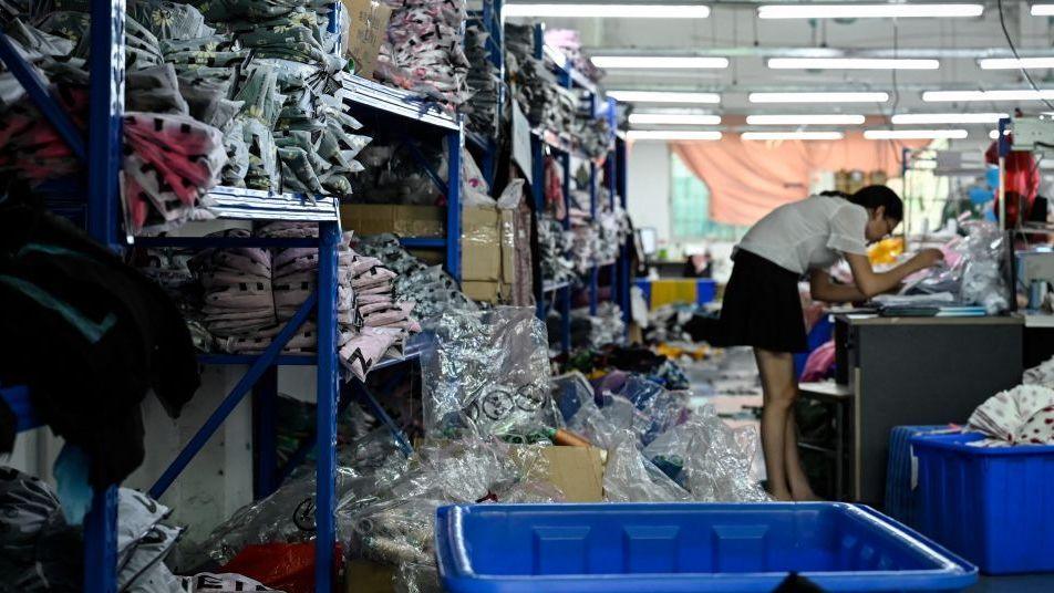พนักงานกำลังผลิตเสื้อผ้าในโรงงานตัดเย็บที่ส่งสินค้าให้ Shein เธอถูกล้อมรอบด้วยกองเสื้อผ้าที่วางอยู่บนชั้นวางสีน้ำเงิน