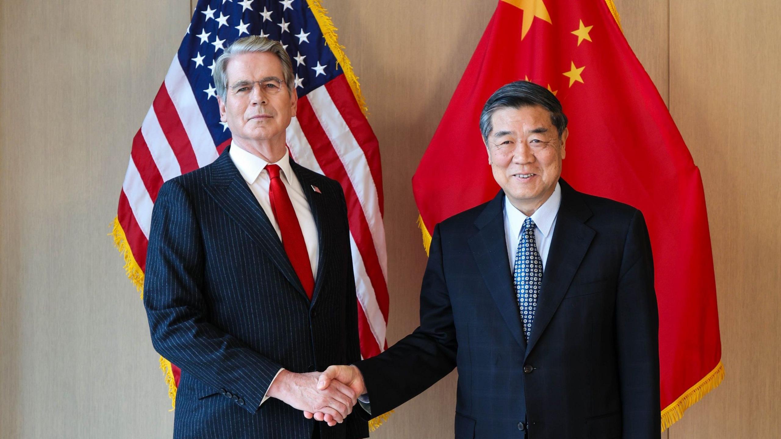 นายสกอตต์ เบสเซนต์ รัฐมนตรีคลังสหรัฐฯ จับมือกับ นายเหอ ลี่เฟิง หัวหน้าผู้เจรจาการค้าของจีน โดยมีธงชาติของทั้งสองประเทศอยู่ด้านหลัง