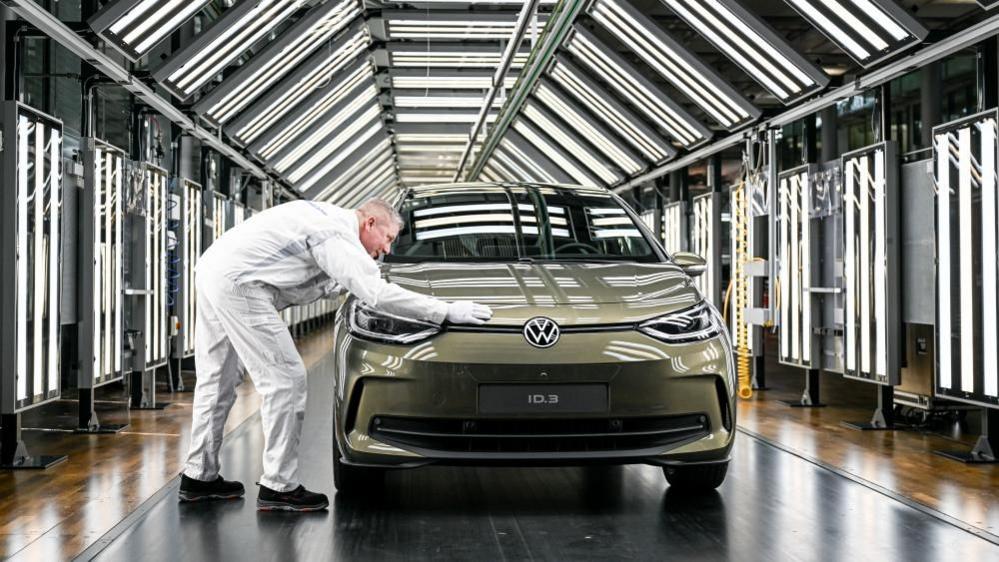 พนักงานของโฟล์คสวาเกนกำลังนำเสนอรถยนต์รุ่น Volkswagen ID.3 โดยเขาวางมือบนรถสีเขียวมอสจอดอยู่ในอุโมงค์ที่เต็มไปด้วยแผงไฟ LED สีขาว