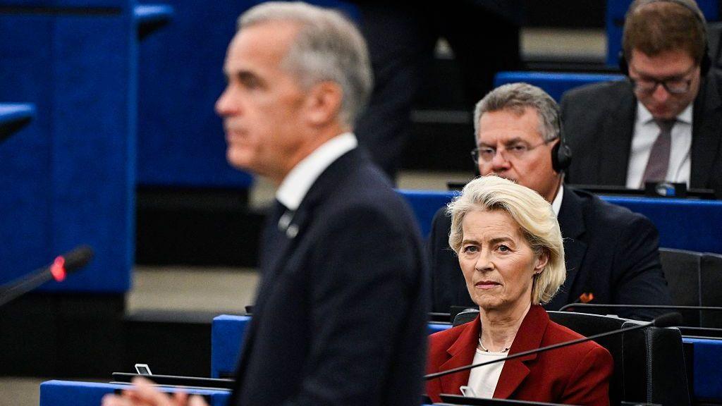 ภาพประธานคณะกรรมาธิการยุโรป Ursula von der Leyen กำลังมอง Mark Carney ขณะกล่าวสุนทรพจน์ต่อรัฐสภายุโรป เธอมีผมบลอนด์สั้นและสวมเสื้อแจ็คเก็ตสีแดงเข้ม