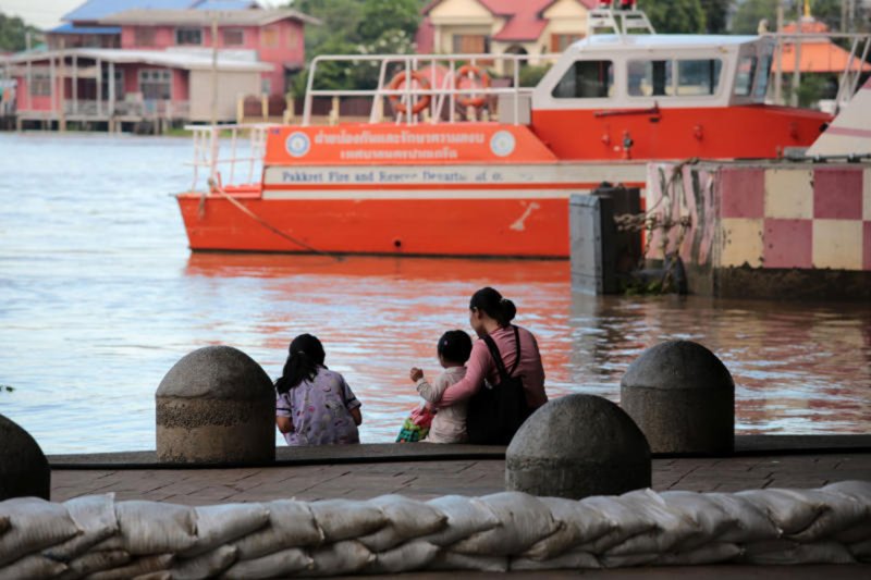 ผู้หญิงและเด็กนั่งอยู่ใกล้ท่าเรือปากเกร็ด จังหวัดนนทบุรี เมื่อวันพฤหัสบดีที่ผ่านมา เจ้าหน้าที่ได้วางกระสอบทรายตามแนวริมฝั่งแม่น้ำเจ้าพระยา หลังจากกรมชลประทานเพิ่มอัตราการระบายน้ำจากเขื่อนเจ้าพระยาในจังหวัดชัยนาท (ภาพ: ชนะท กตัญญู)