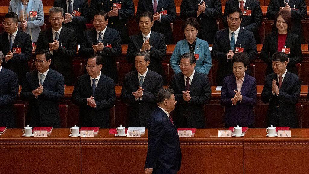 แถวของเจ้าหน้าที่ยืนปรบมือภายในห้องประชุมสภานิติบัญญัติขนาดใหญ่ที่มีที่นั่งแบบขั้นบันไดและโต๊ะไม้ขัดเงา ด้านหน้า ประธานาธิบดีสี จิ้นผิง เดินผ่านแถวหน้าสุด ขณะที่ผู้เข้าร่วมด้านหลังยังคงยืนและปรบมือ