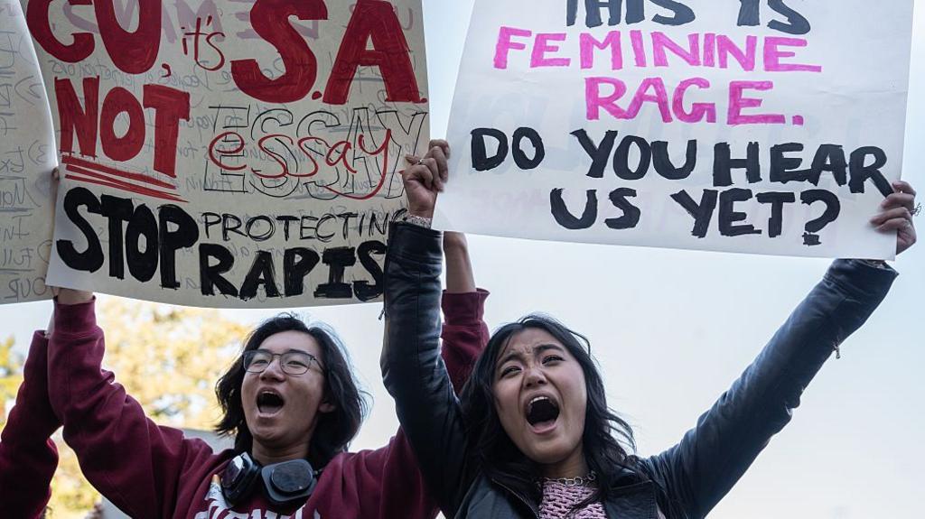 ผู้ประท้วงสองคนถือป้าย โดยป้ายหนึ่งเขียนว่า "CU, it's S.A. not essay. Stop protecting rapists" และอีกป้ายเขียนว่า "This is feminine rage. Do you hear us yet?"