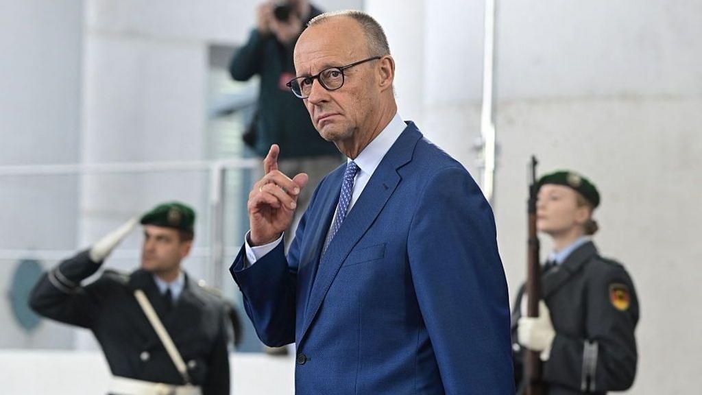 นายกรัฐมนตรีเยอรมนี Friedrich Merz สวมชุดสูทสีน้ำเงิน ยกมือขึ้น มีนายทหารสองนายยืนอยู่ด้านหลังในภาพเบลอ