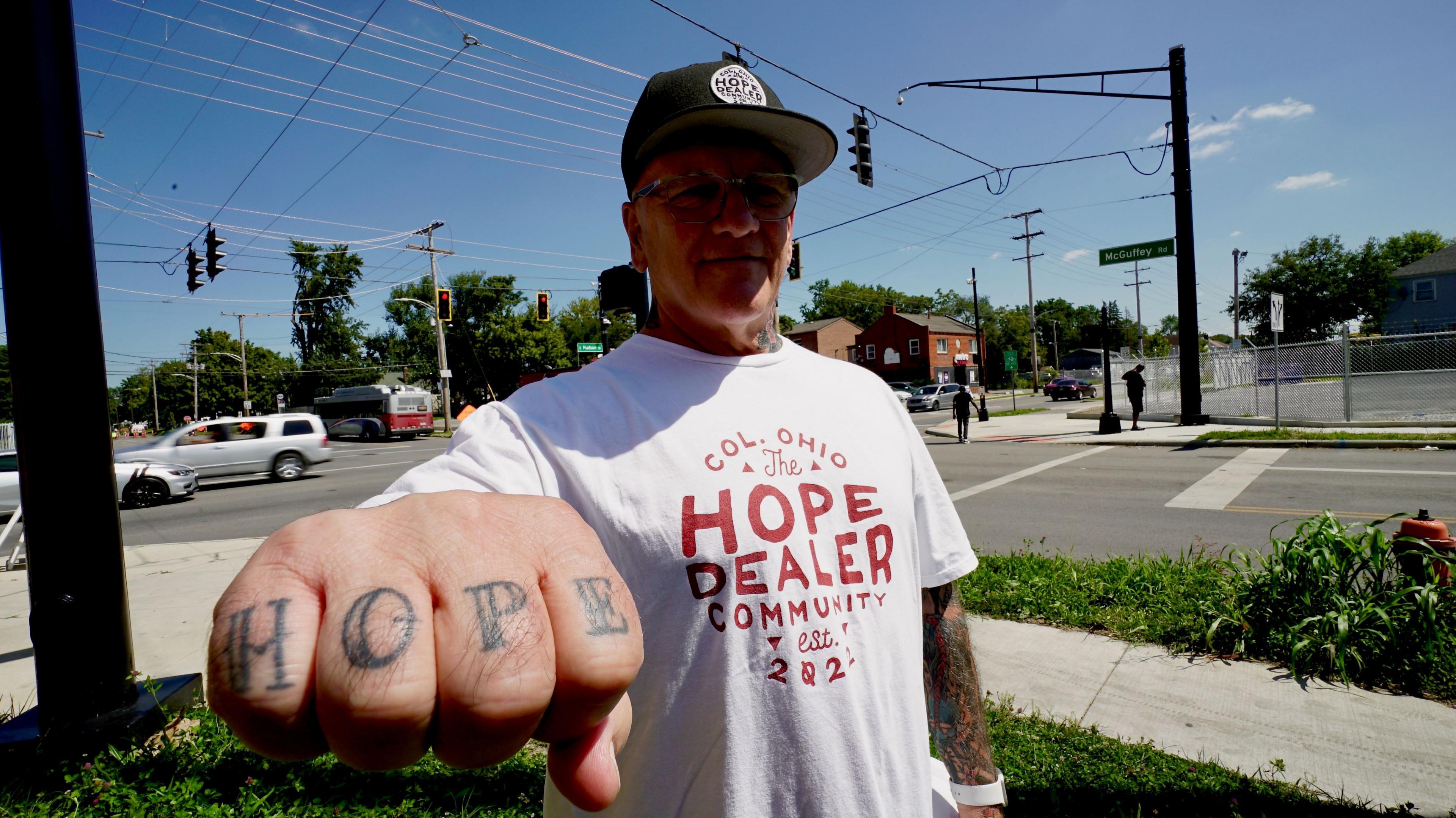 สกอตต์ แซนเดอร์ส วัย 65 ปี สวมหมวกเบสบอลและเสื้อยืดที่เขียนว่า The Hope Dealer Community เขากำหมัดชี้มาที่กล้อง และที่สนับมือของเขามีคำว่า Hope